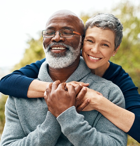  Woman in blue shirt hugging man in gray sweater from behind by a tree outside