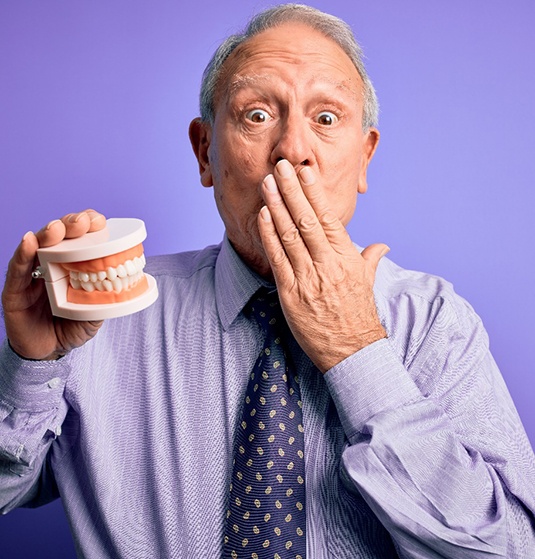 Older man in purple shirt and tie holding false teeth in one hand and convering his mouth with the other