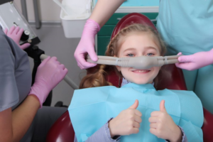 Child giving two thumbs up as dentists administer laughing gas